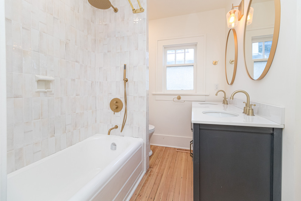 Small transitional white tile medium tone wood floor, beige floor and double-sink drop-in bathtub photo with shaker cabinets, blue cabinets, a two-piece toilet, white walls, an undermount sink, soapstone countertops, white countertops and a freestanding vanity