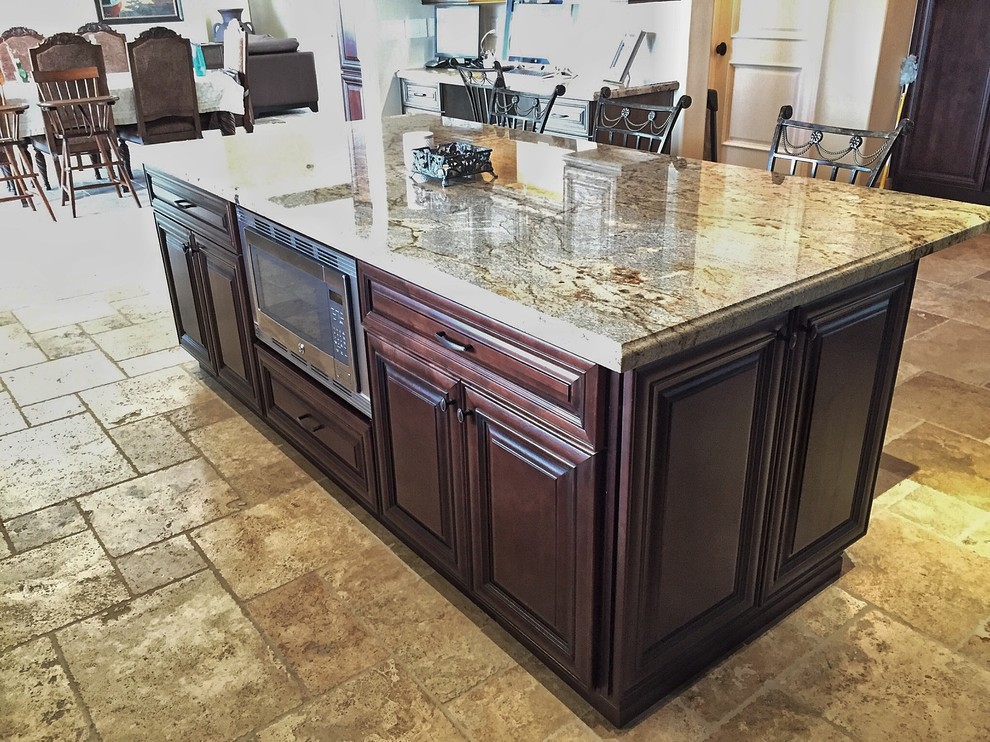 Photo of a large traditional u-shaped eat-in kitchen in Phoenix with a farmhouse sink, raised-panel cabinets, dark wood cabinets, granite benchtops, beige splashback, travertine splashback, stainless steel appliances, travertine floors, with island, beige floor and beige benchtop.