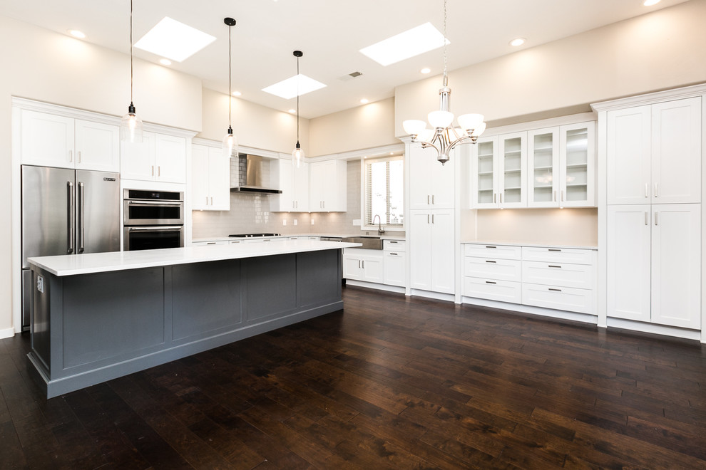 Large transitional l-shaped dark wood floor and brown floor eat-in kitchen photo in Albuquerque with a farmhouse sink, shaker cabinets, white cabinets, quartz countertops, gray backsplash, subway tile backsplash, stainless steel appliances and an island