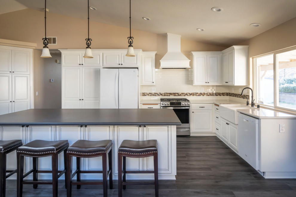 This is an example of a large traditional l-shaped kitchen/diner in Los Angeles with a belfast sink, raised-panel cabinets, white cabinets, engineered stone countertops, white splashback, cement tile splashback, white appliances, vinyl flooring, an island, grey floors and black worktops.