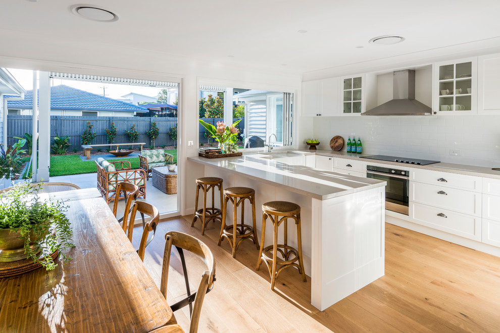 Photo of a mid-sized beach style u-shaped eat-in kitchen in Gold Coast - Tweed with a farmhouse sink, shaker cabinets, white cabinets, quartz benchtops, white splashback, ceramic splashback, stainless steel appliances, light hardwood floors, with island and brown floor.