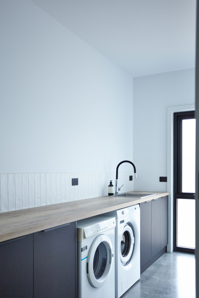 Inspiration for a mid-sized single-wall dedicated laundry room in Other with a single-bowl sink, grey cabinets, wood benchtops, white splashback, subway tile splashback, concrete floors, a side-by-side washer and dryer and grey floor.