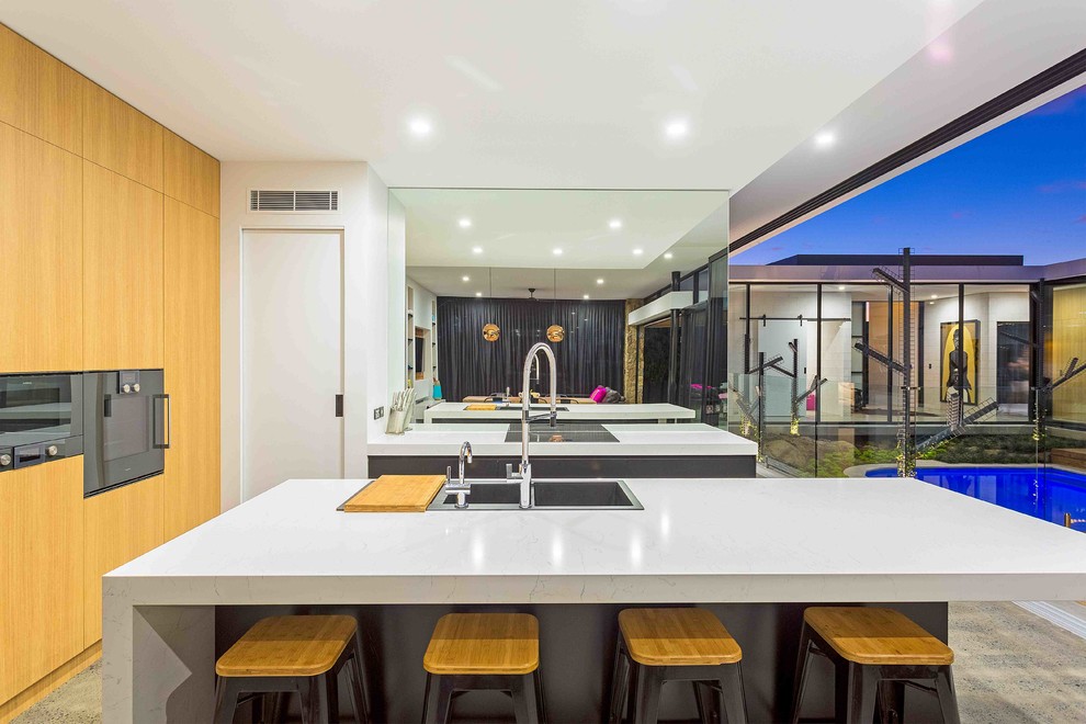 Inspiration for a contemporary galley open plan kitchen in Gold Coast - Tweed with a drop-in sink, flat-panel cabinets, metallic splashback, mirror splashback, black appliances, concrete floors, with island, grey floor, light wood cabinets, granite benchtops and white benchtop.