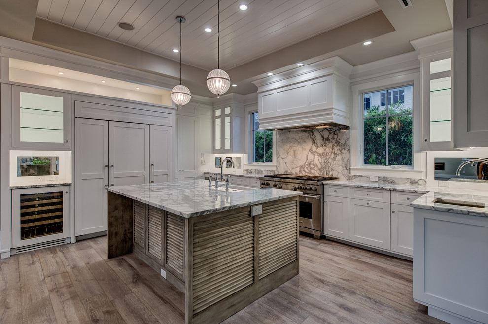 Large transitional l-shaped open plan kitchen in Other with a farmhouse sink, recessed-panel cabinets, white cabinets, marble benchtops, mirror splashback, stainless steel appliances, light hardwood floors and with island.