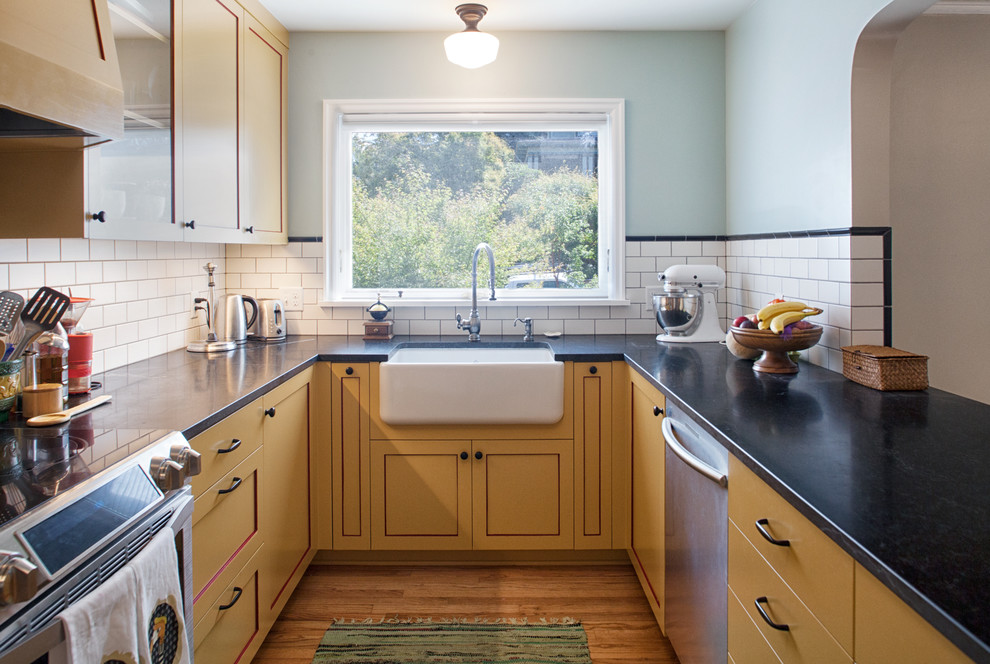 Design ideas for a small traditional u-shaped kitchen in Portland with a farmhouse sink, shaker cabinets, yellow cabinets, soapstone benchtops, white splashback, subway tile splashback, stainless steel appliances, light hardwood floors and no island.