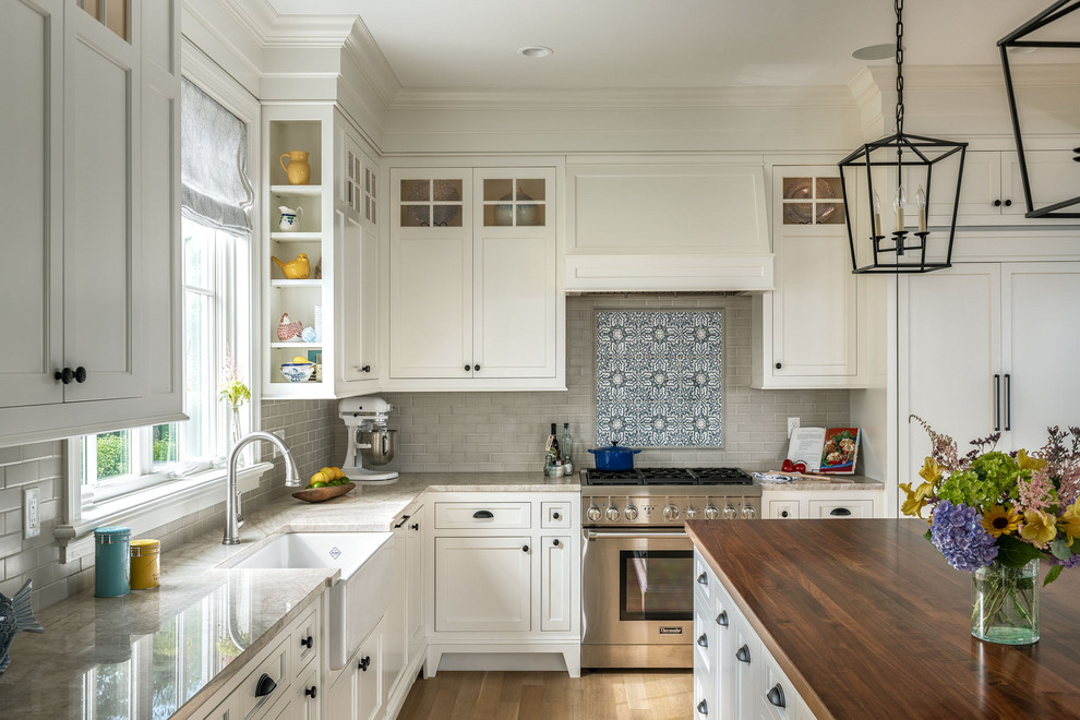 Photo of a beach style l-shaped kitchen in Boston with a farmhouse sink, recessed-panel cabinets, white cabinets, grey splashback, subway tile splashback, stainless steel appliances, medium hardwood floors, with island, brown floor and beige benchtop.