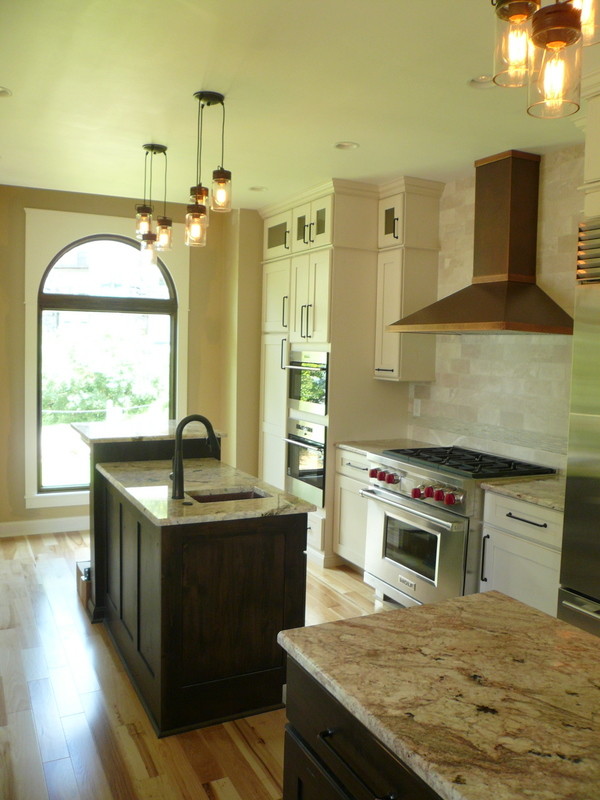 Example of a mid-sized mountain style galley light wood floor eat-in kitchen design with a farmhouse sink, shaker cabinets, white cabinets, granite countertops, beige backsplash, stone tile backsplash, stainless steel appliances and two islands