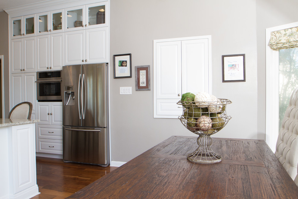 Eat-in kitchen - large transitional l-shaped medium tone wood floor eat-in kitchen idea in Austin with a farmhouse sink, raised-panel cabinets, white cabinets, granite countertops, beige backsplash, ceramic backsplash, stainless steel appliances and an island