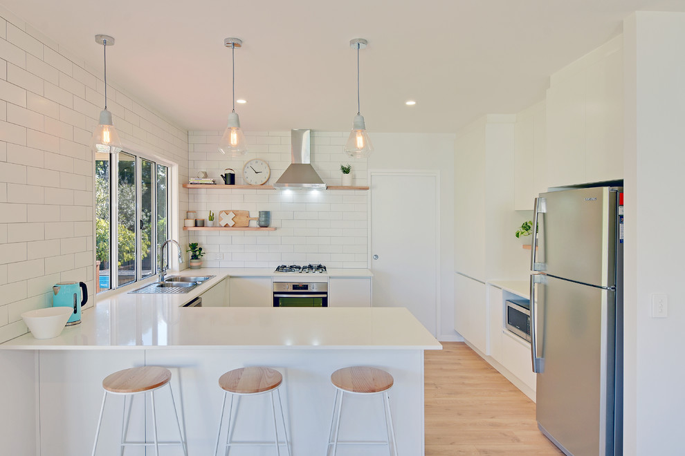 Design ideas for a beach style u-shaped separate kitchen in Sunshine Coast with a drop-in sink, flat-panel cabinets, white cabinets, white splashback, subway tile splashback, stainless steel appliances, light hardwood floors, a peninsula and beige floor.