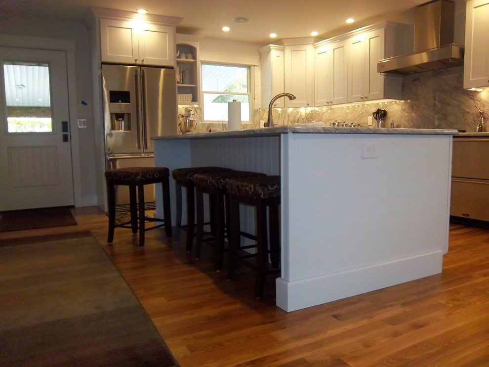 Example of a mid-sized transitional l-shaped medium tone wood floor and brown floor open concept kitchen design with an undermount sink, recessed-panel cabinets, white cabinets, granite countertops, gray backsplash, stone slab backsplash, stainless steel appliances and an island
