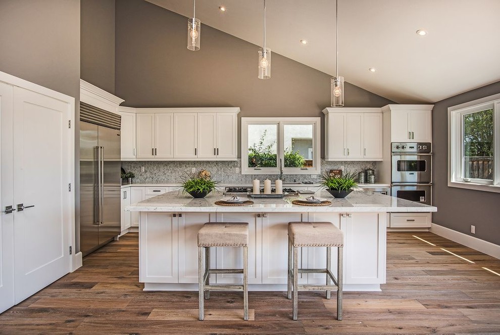 Inspiration for a large transitional l-shaped medium tone wood floor and brown floor open concept kitchen remodel in Orange County with an undermount sink, shaker cabinets, white cabinets, marble countertops, gray backsplash, glass tile backsplash, stainless steel appliances and an island
