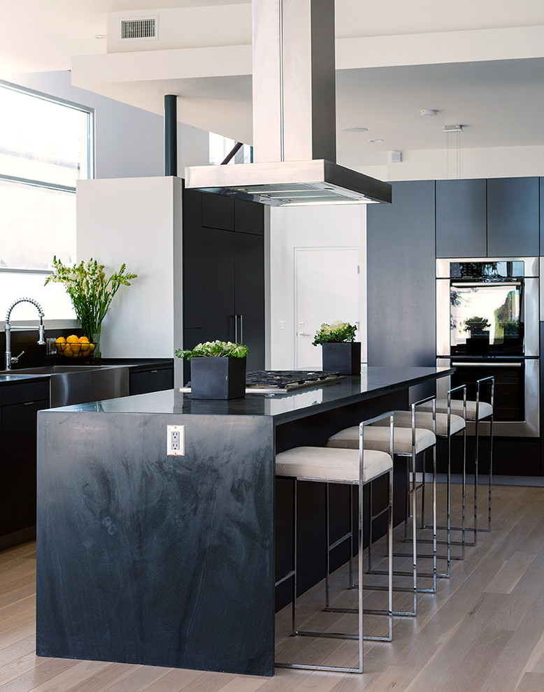 Inspiration for a large contemporary u-shaped eat-in kitchen in Los Angeles with a farmhouse sink, flat-panel cabinets, stainless steel appliances, light hardwood floors, with island, wood benchtops, window splashback, beige floor and black benchtop.