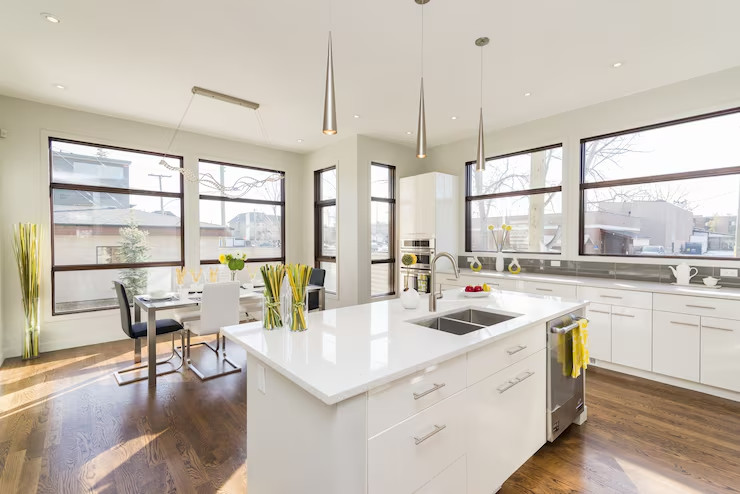 Sunlit contemporary kitchen and dining area with a white island, pendant lights, wood floors, and large wraparound windows.