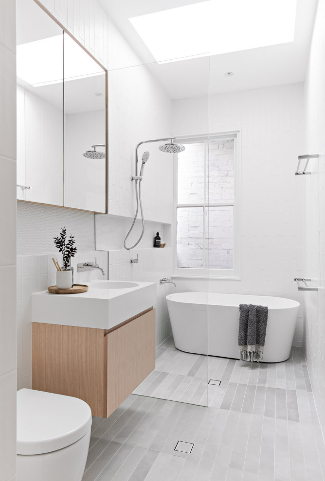 Photo of a mid-sized modern kids bathroom in Sydney with light wood cabinets, a freestanding tub, white tile, ceramic tile, white walls, porcelain floors, solid surface benchtops, grey floor, an open shower, white benchtops, a niche, a single vanity, a floating vanity, flat-panel cabinets, a curbless shower, a one-piece toilet and an integrated sink.