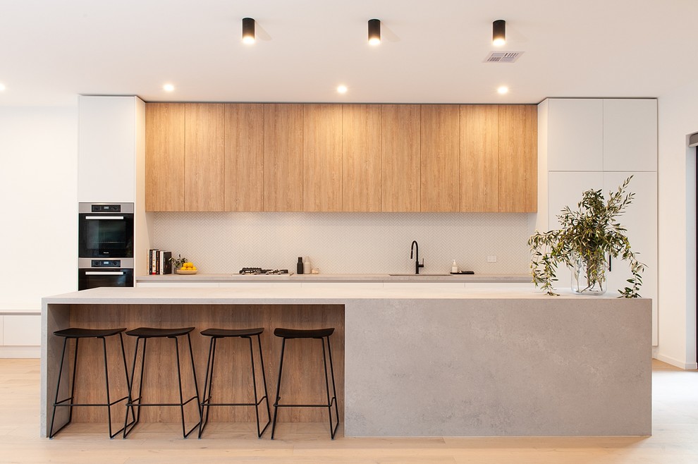 Photo of an expansive contemporary galley open plan kitchen in Melbourne with an integrated sink, quartz benchtops, grey splashback, marble splashback, black appliances, light hardwood floors, with island, grey benchtop, flat-panel cabinets, medium wood cabinets and brown floor.