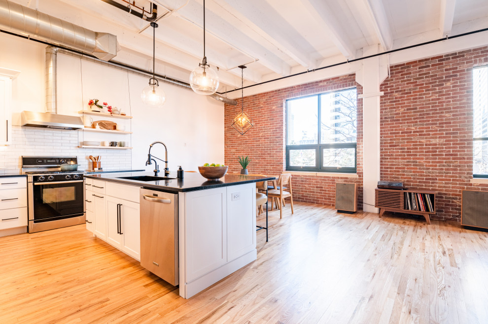 Large industrial l-shaped eat-in kitchen in Denver with an integrated sink, shaker cabinets, white cabinets, soapstone benchtops, white splashback, ceramic splashback, stainless steel appliances, light hardwood floors, with island, black benchtop and exposed beam.