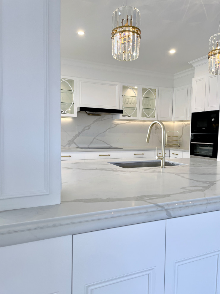 Large traditional l-shaped open plan kitchen in Sydney with an undermount sink, recessed-panel cabinets, white cabinets, quartz benchtops, white splashback, engineered quartz splashback, black appliances, light hardwood floors, with island, brown floor and white benchtop.