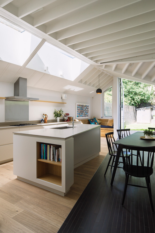 This is an example of a mid-sized contemporary eat-in kitchen in London with a drop-in sink, flat-panel cabinets, white cabinets, solid surface benchtops, grey splashback, light hardwood floors, with island, brown floor and metal splashback.