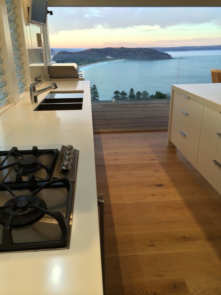 Design ideas for a large beach style galley open plan kitchen in Sydney with a double-bowl sink, white cabinets, solid surface benchtops, white splashback, glass sheet splashback, stainless steel appliances, light hardwood floors and with island.