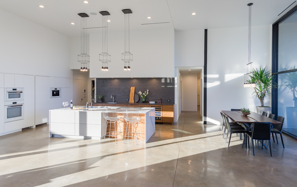 Photo of a mid-sized modern l-shaped open plan kitchen in Oklahoma City with an undermount sink, flat-panel cabinets, quartz benchtops, black splashback, white appliances, concrete floors and with island.