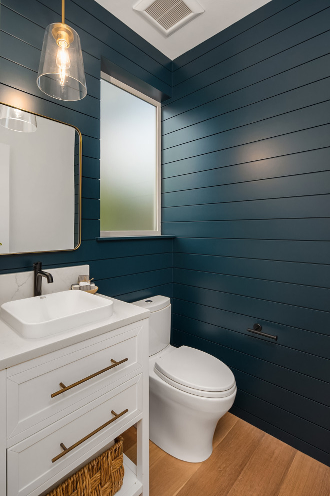 Photo of a small beach style cloakroom in Seattle with recessed-panel cabinets, white cabinets, a two-piece toilet, blue walls, light hardwood flooring, a built-in sink, quartz worktops, white worktops, a freestanding vanity unit and tongue and groove walls.