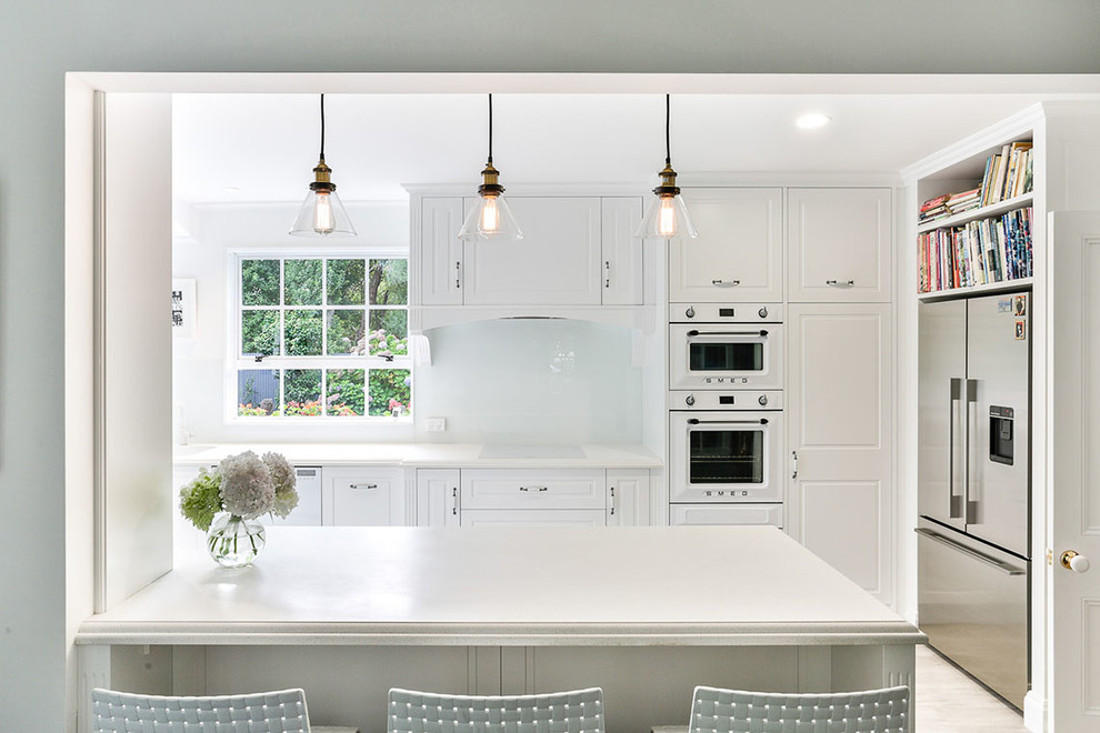 Photo of a mid-sized country u-shaped eat-in kitchen in Other with an undermount sink, recessed-panel cabinets, white cabinets, solid surface benchtops, white splashback, glass sheet splashback, white appliances, vinyl floors, no island, brown floor and white benchtop.