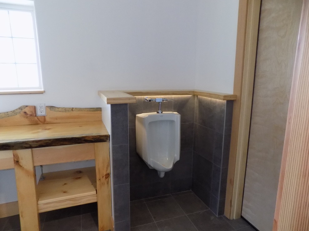 This is an example of a mid-sized industrial 3/4 bathroom in Boston with furniture-like cabinets, light wood cabinets, an open shower, an urinal, white walls, pebble tile floors, a drop-in sink, wood benchtops, grey floor, a sliding shower screen and brown benchtops.