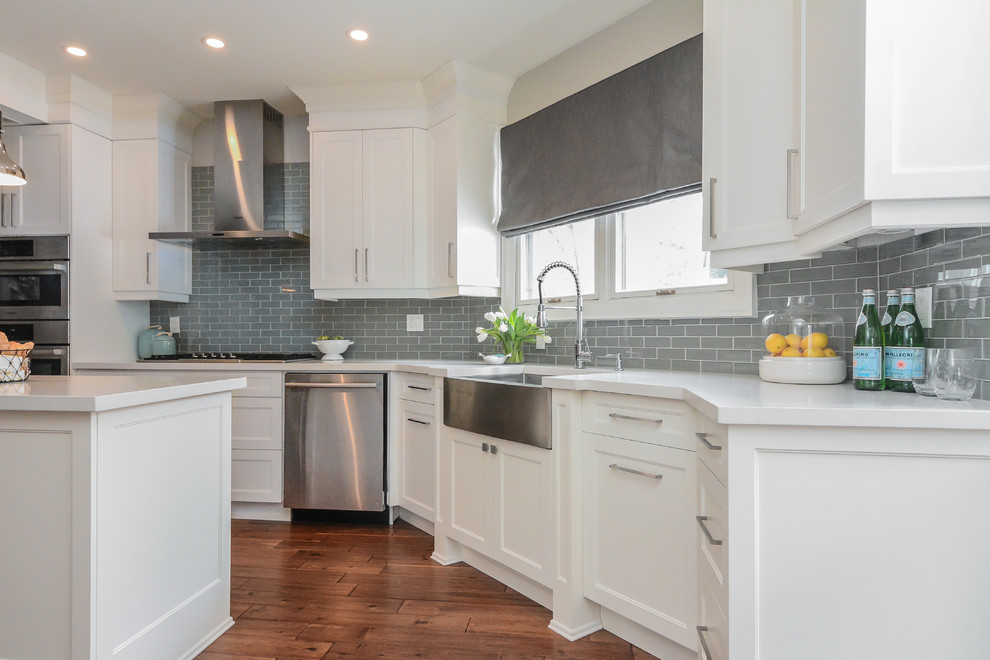 This is an example of a large transitional l-shaped eat-in kitchen in Toronto with a farmhouse sink, raised-panel cabinets, white cabinets, quartz benchtops, grey splashback, glass tile splashback, stainless steel appliances, medium hardwood floors, with island and brown floor.
