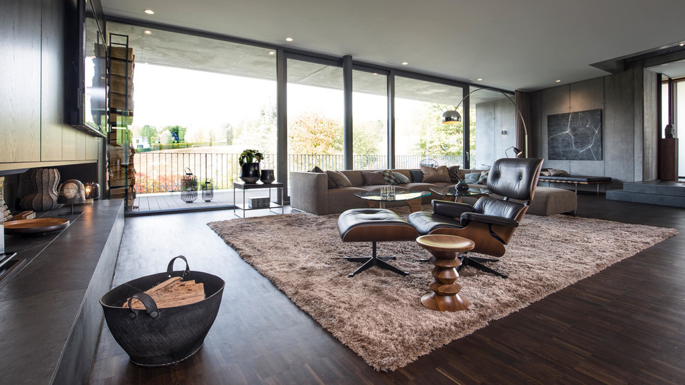 This is an example of a large contemporary open concept family room in Dortmund with grey walls, dark hardwood floors, a two-sided fireplace, a wood fireplace surround, a wall-mounted tv and brown floor.