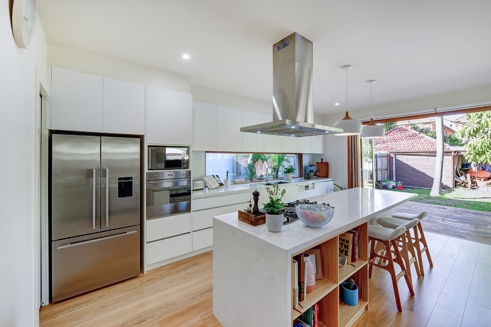 Photo of a large scandinavian galley open plan kitchen in Sydney with an undermount sink, flat-panel cabinets, white cabinets, quartz benchtops, white splashback, glass sheet splashback, stainless steel appliances, light hardwood floors, with island, brown floor and white benchtop.