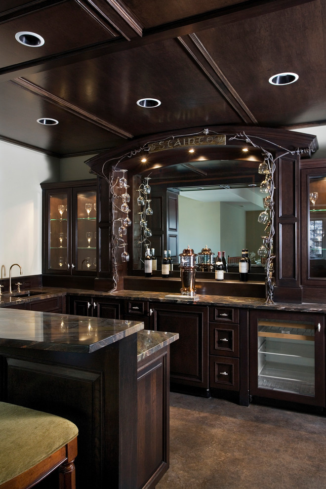 This is an example of an expansive traditional u-shaped seated home bar in Chicago with an undermount sink, recessed-panel cabinets, dark wood cabinets, granite benchtops, mirror splashback, concrete floors and brown floor.