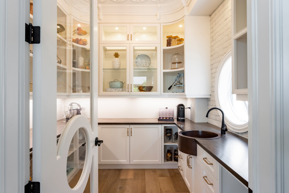 This is an example of a transitional u-shaped kitchen pantry in Other with a farmhouse sink, recessed-panel cabinets, white cabinets, wood benchtops, white splashback, brick splashback, medium hardwood floors, no island, brown floor and brown benchtop.
