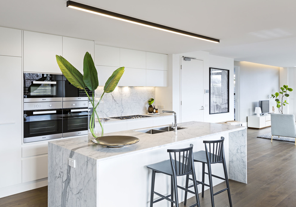 This is an example of a mid-sized contemporary galley kitchen in Melbourne with flat-panel cabinets, white cabinets, marble benchtops, stone slab splashback, stainless steel appliances, medium hardwood floors, with island, brown floor, white benchtop, a double-bowl sink and white splashback.