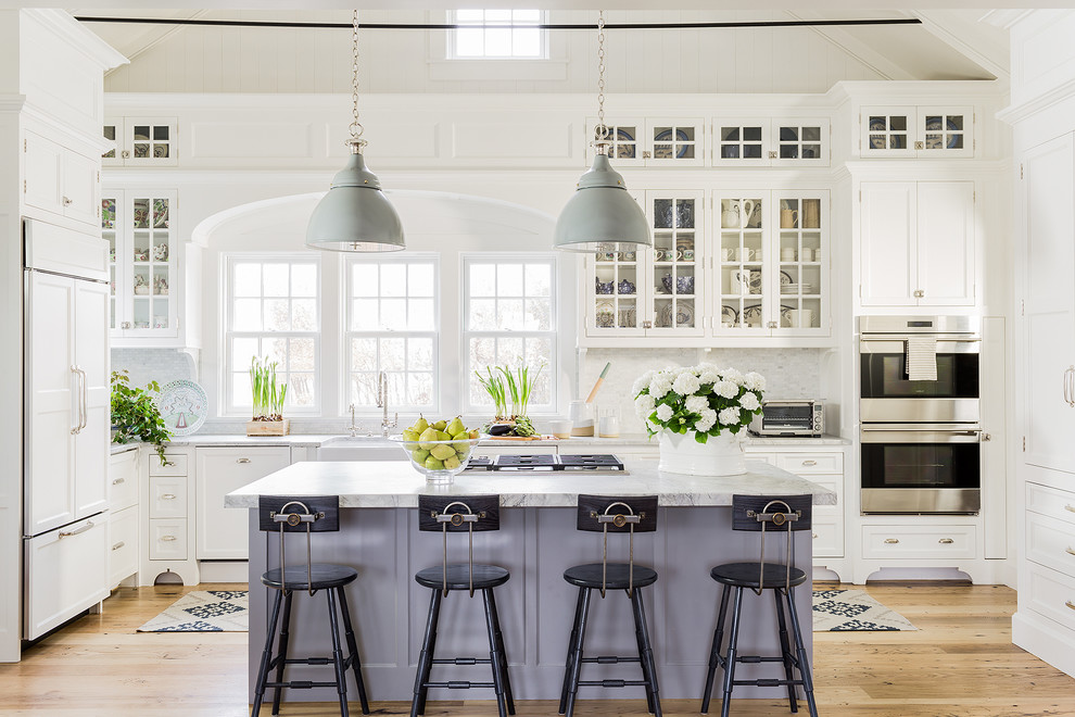 Large traditional kitchen in Boston with a farmhouse sink, white cabinets, quartzite benchtops, white splashback, panelled appliances, light hardwood floors, with island and glass-front cabinets.