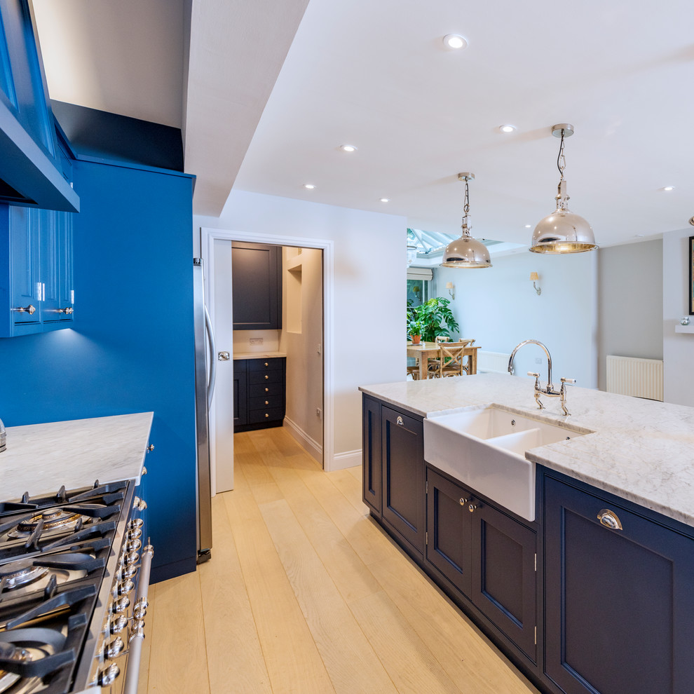 This is an example of a large modern galley open plan kitchen in Other with a farmhouse sink, recessed-panel cabinets, blue cabinets, marble benchtops, multi-coloured splashback, stainless steel appliances, light hardwood floors, with island, white floor and white benchtop.