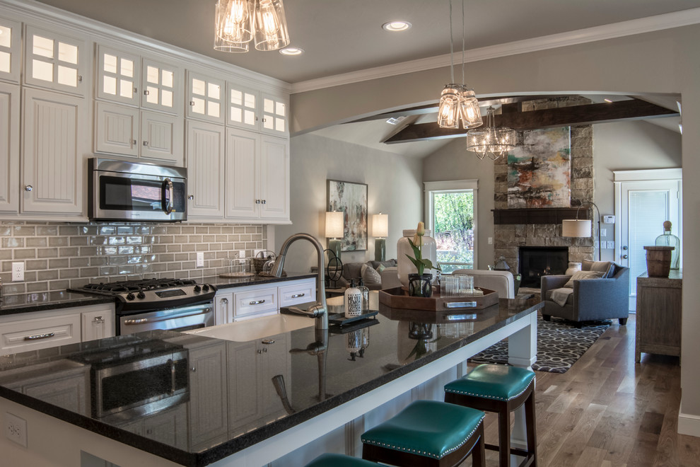 Inspiration for a mid-sized arts and crafts eat-in kitchen in Oklahoma City with a farmhouse sink, glass-front cabinets, white cabinets, granite benchtops, beige splashback, stainless steel appliances, light hardwood floors and with island.