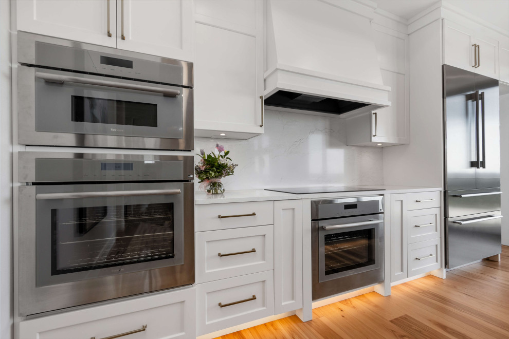 Example of a mid-sized transitional single-wall medium tone wood floor, brown floor and tray ceiling eat-in kitchen design with an undermount sink, shaker cabinets, white cabinets, quartz countertops, white backsplash, quartz backsplash, stainless steel appliances, an island and white countertops