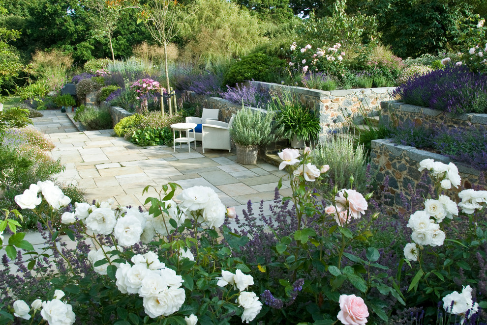 Large country backyard full sun garden in Channel Islands with a retaining wall and natural stone pavers for summer.