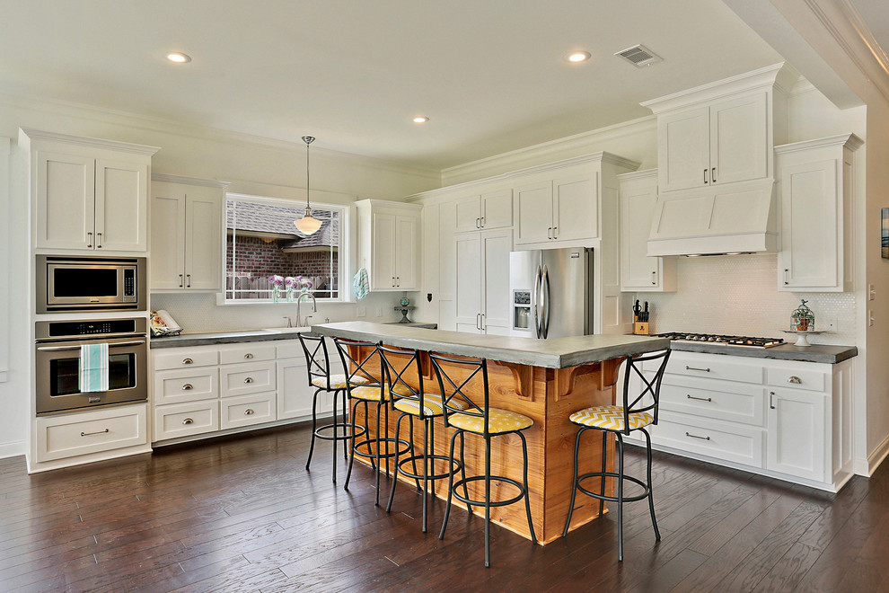 Large beach style l-shaped open plan kitchen in New Orleans with a double-bowl sink, shaker cabinets, white cabinets, concrete benchtops, white splashback, ceramic splashback, stainless steel appliances, dark hardwood floors and with island.