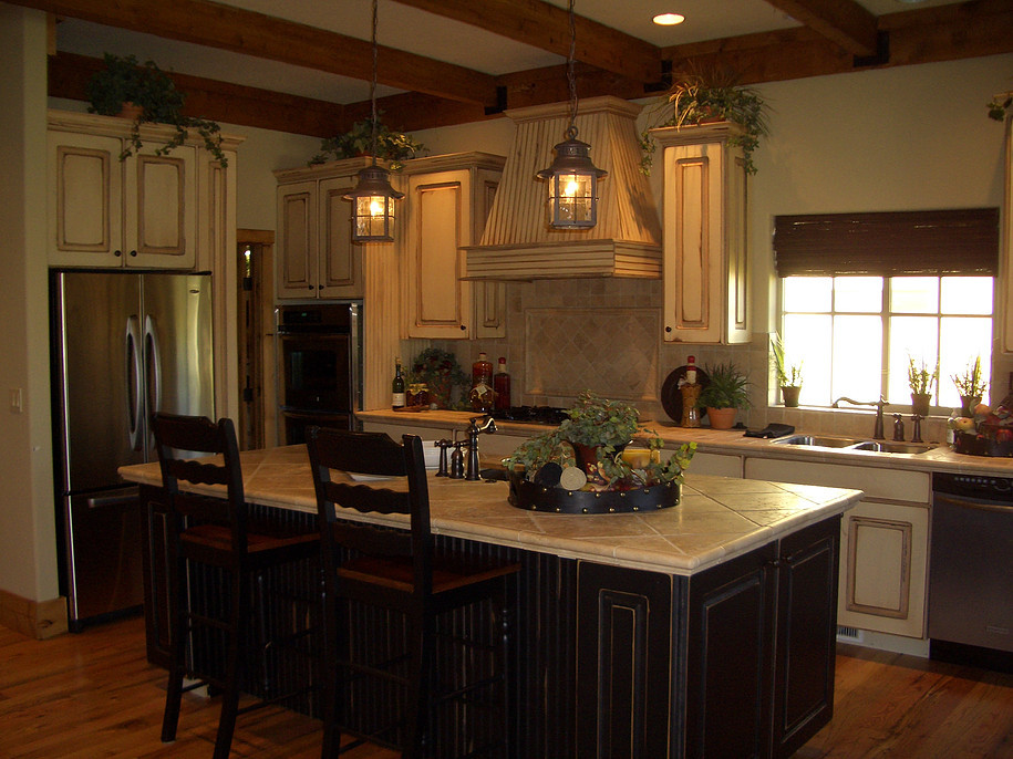 Mid-sized country l-shaped eat-in kitchen in San Francisco with an undermount sink, raised-panel cabinets, distressed cabinets, tile benchtops, beige splashback, stone tile splashback, stainless steel appliances, medium hardwood floors, with island, brown floor and beige benchtop.