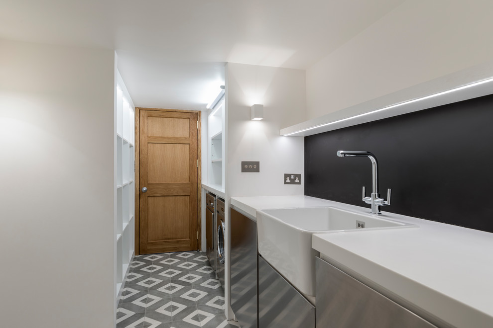 Design ideas for a transitional galley utility room in London with a farmhouse sink, flat-panel cabinets, stainless steel cabinets, solid surface benchtops, white walls, porcelain floors, a side-by-side washer and dryer and grey floor.