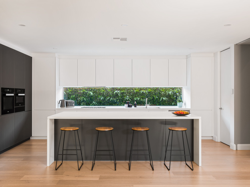 Inspiration for a large modern l-shaped open plan kitchen in Sydney with a double-bowl sink, open cabinets, white cabinets, solid surface benchtops, white splashback, window splashback, black appliances, light hardwood floors, with island, brown floor and white benchtop.