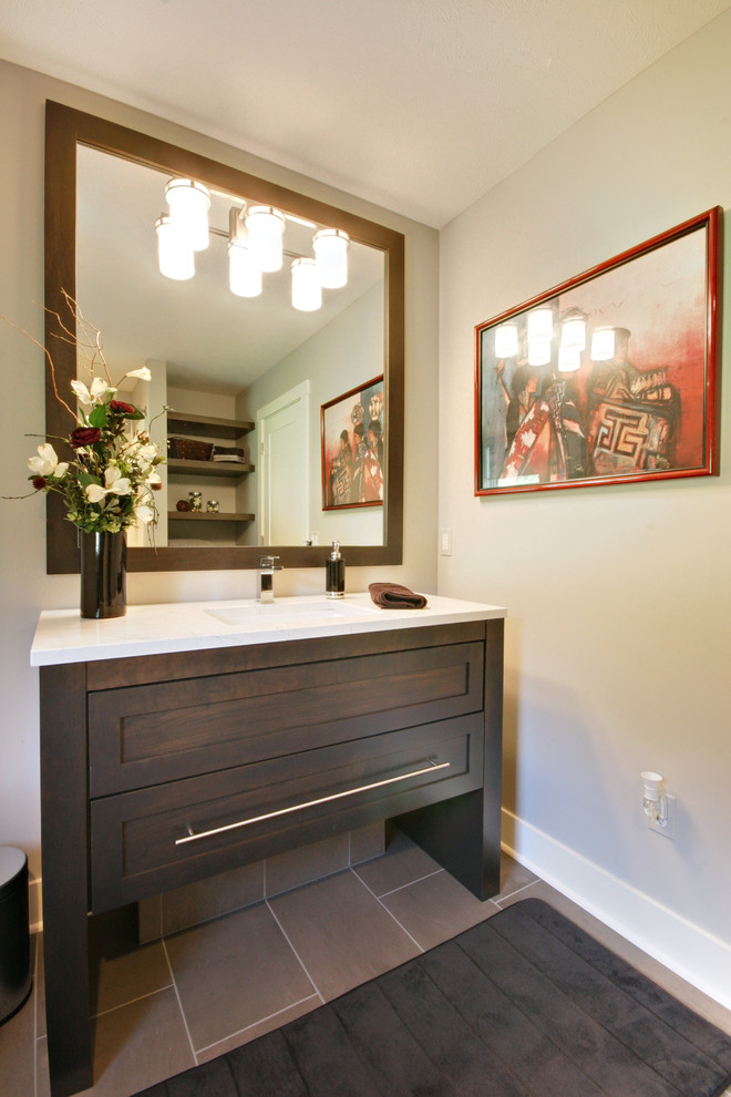This is an example of a mid-sized contemporary powder room in Grand Rapids with recessed-panel cabinets, dark wood cabinets, a one-piece toilet, black tile, porcelain tile, grey walls, porcelain floors, a drop-in sink, engineered quartz benchtops, grey floor and white benchtops.