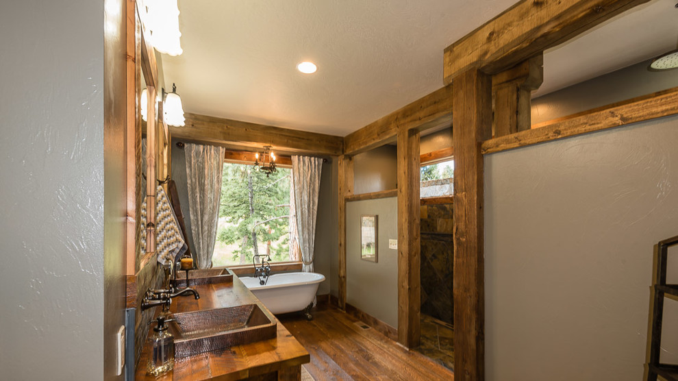 Example of a large mountain style master stone tile medium tone wood floor and brown floor claw-foot bathtub design with medium tone wood cabinets, gray walls, a vessel sink and wood countertops