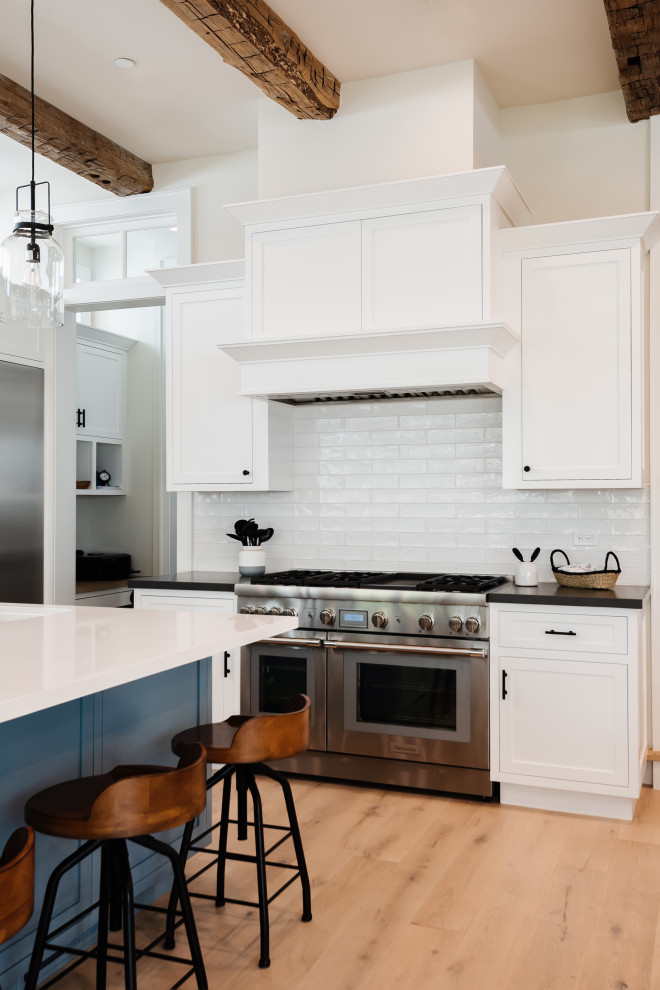 Large country u-shaped open plan kitchen in San Diego with a farmhouse sink, shaker cabinets, white cabinets, quartz benchtops, white splashback, porcelain splashback, stainless steel appliances, light hardwood floors, with island, brown floor, black benchtop and exposed beam.