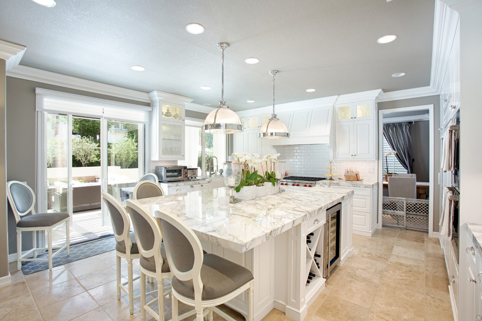 This is an example of a large transitional l-shaped eat-in kitchen with a farmhouse sink, raised-panel cabinets, white cabinets, marble benchtops, white splashback, subway tile splashback, stainless steel appliances, travertine floors, with island and beige floor.