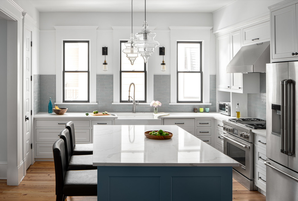 Inspiration for an expansive transitional l-shaped kitchen in Atlanta with a farmhouse sink, shaker cabinets, white cabinets, quartz benchtops, grey splashback, subway tile splashback, stainless steel appliances, with island, light hardwood floors and white benchtop.