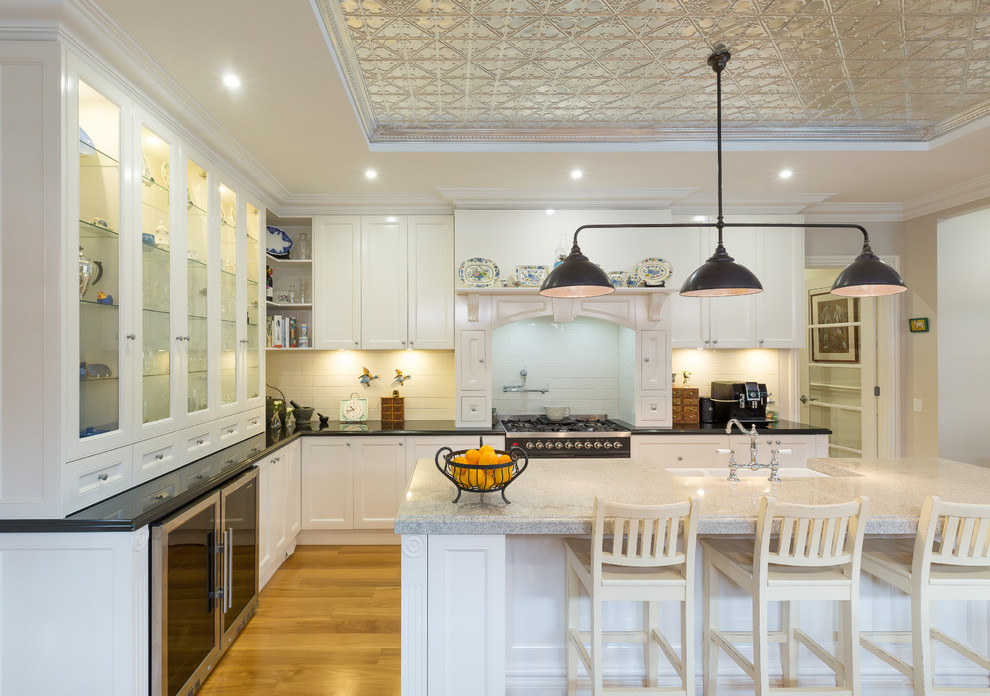 This is an example of a mid-sized traditional l-shaped eat-in kitchen in Melbourne with an undermount sink, white cabinets, white splashback, ceramic splashback, stainless steel appliances, light hardwood floors, with island and brown floor.