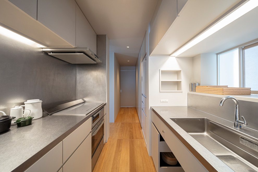 Photo of a small modern galley separate kitchen in Tokyo with an integrated sink, white cabinets, stainless steel benchtops, stainless steel appliances, medium hardwood floors, no island and white benchtop.