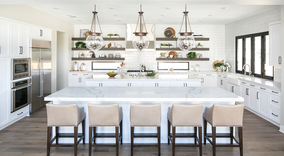 Photo of a beach style kitchen in San Diego with white cabinets, white splashback, subway tile splashback, medium hardwood floors, multiple islands, a farmhouse sink, shaker cabinets, stainless steel appliances and white benchtop.
